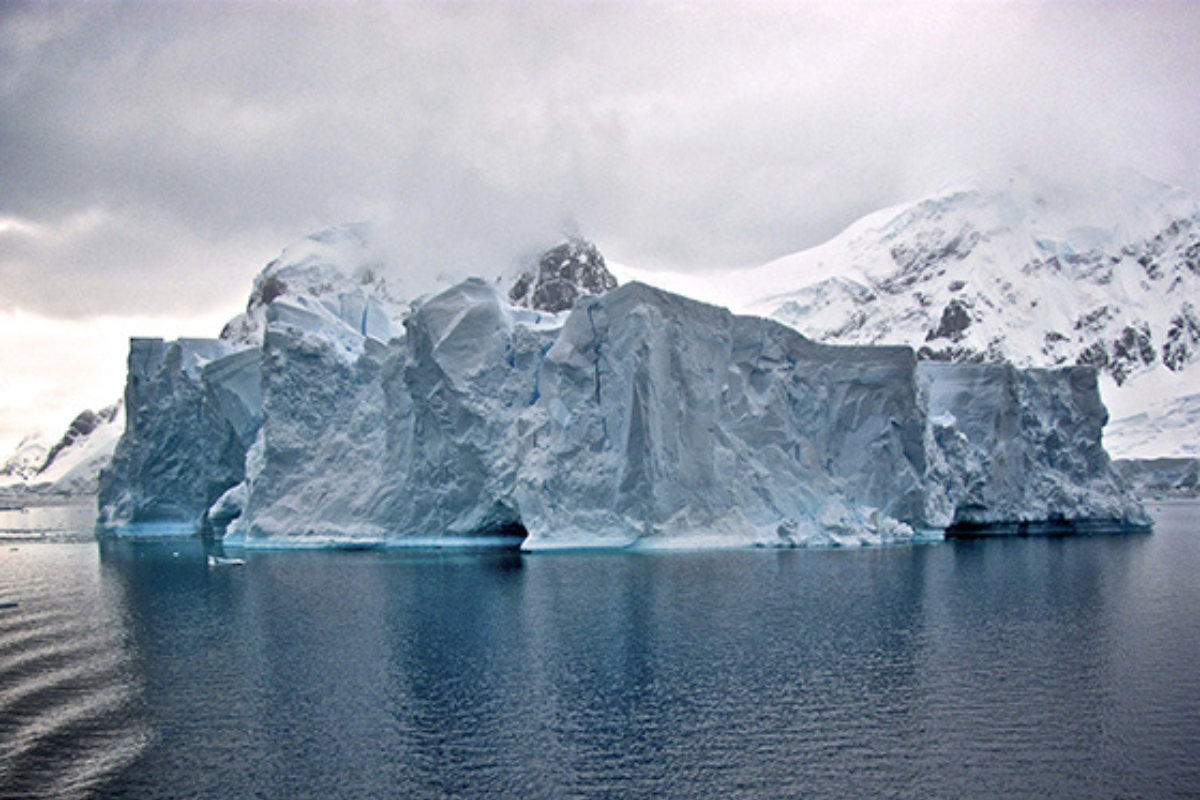 A large iceberg floating in the ocean with snow-covered mountains in the background, under a cloudy sky.